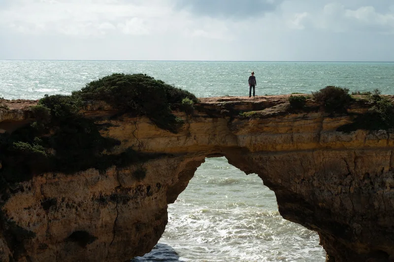 A small figure on a coastal rock arch gives scale to the sea and stone.