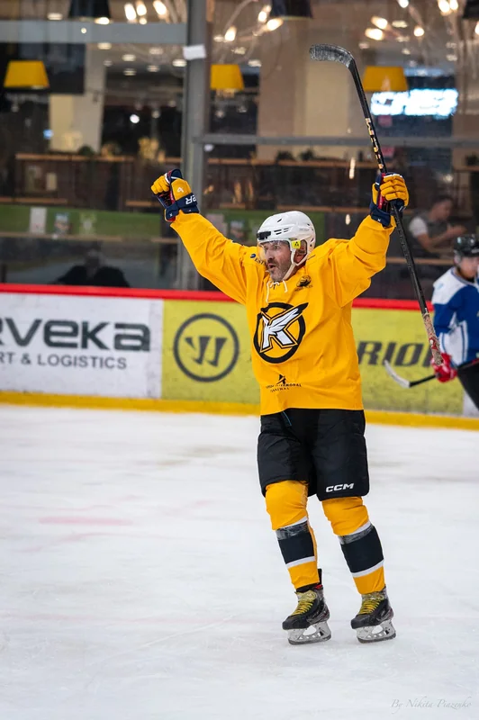 A yellow-shirted hockey player raises both arms and his stick.