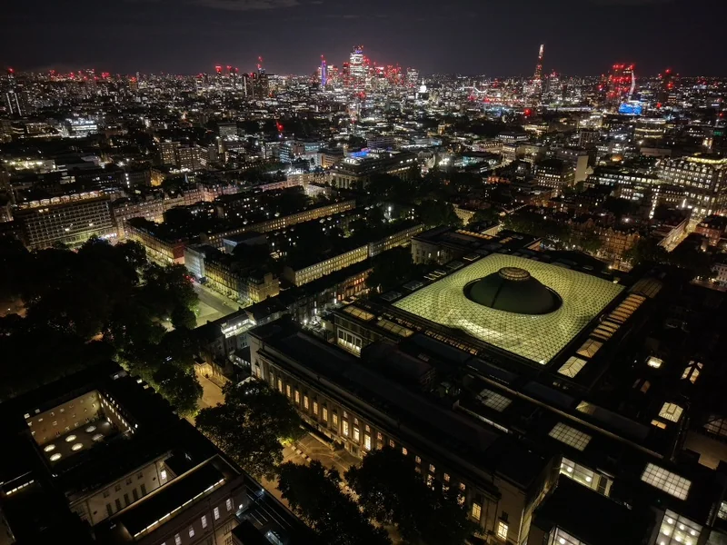 Aerial night view across a glass-roofed courtyard towards the city skyline