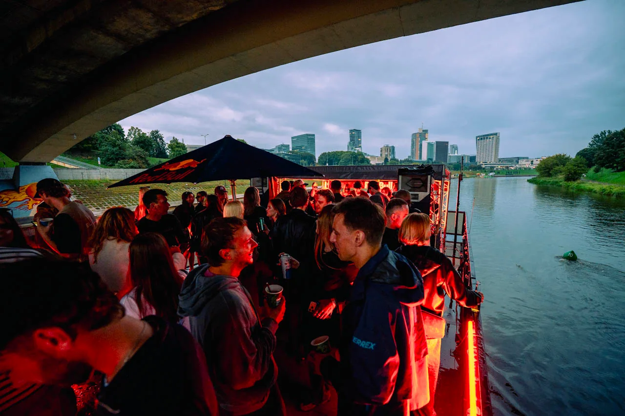 Crowd and DJ lights aboard the District boat beneath a Vilnius bridge at dusk