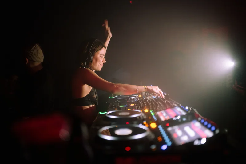 DJ raising one hand beside a bright spotlight at E1