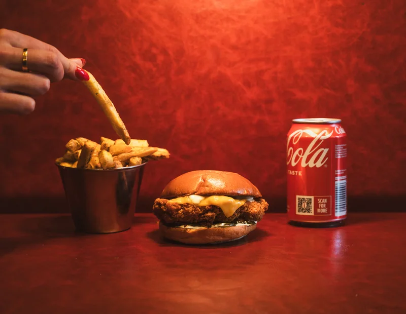 Hand lifting a fry beside a chicken cheeseburger and Coca-Cola against a red background