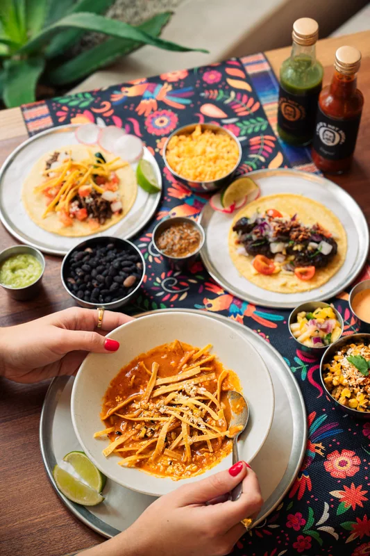 Hands around a table of tacos, soup and sides