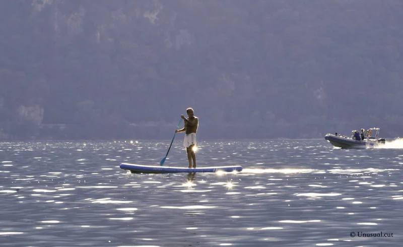 Paddleboarder crossing sunlit water with a motorboat and mountains behind