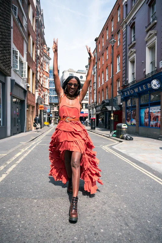 Woman in an orange dress raising her arms on a Soho street