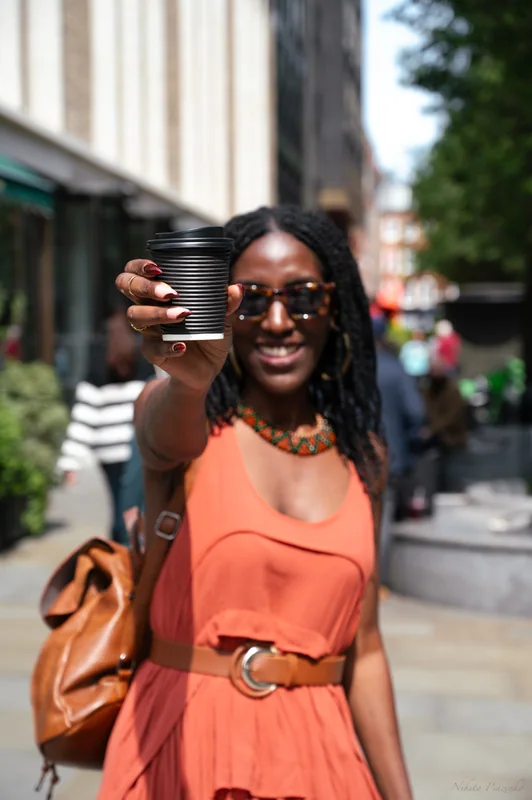 Woman in an orange dress offering a coffee towards the camera on a Soho street